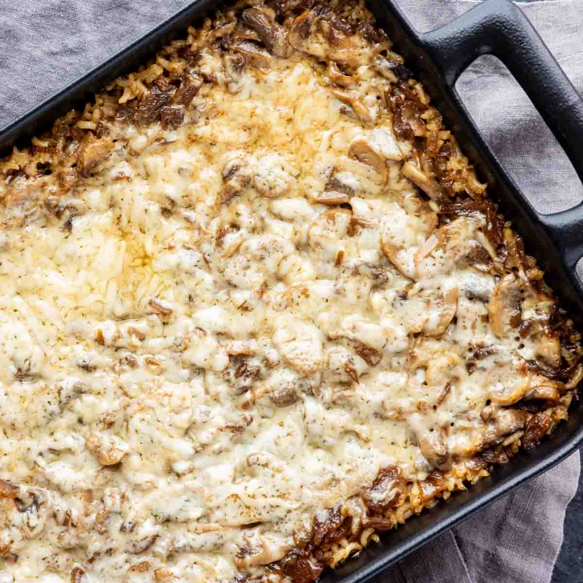 Mushroom and stick of butter rice casserole topped with melted cheese in a black rectangular baking dish.