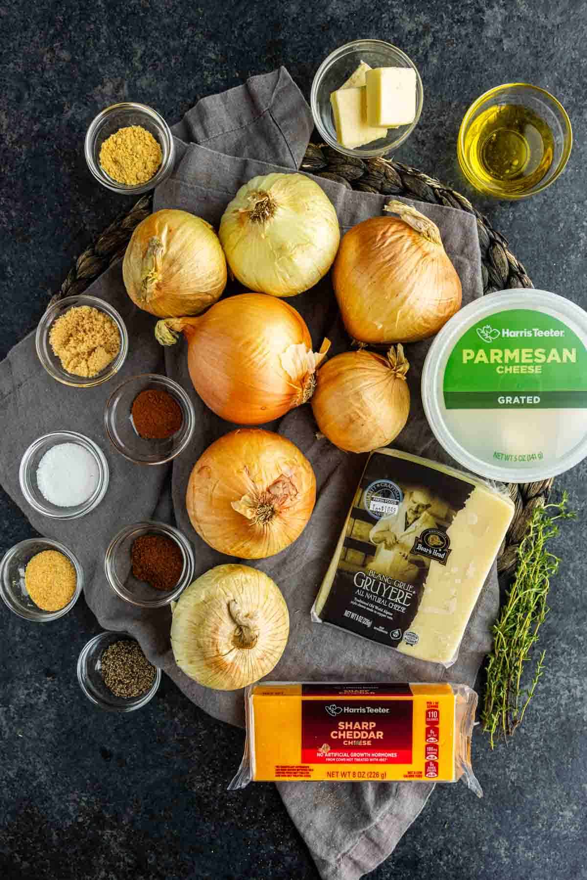 Ingredients for a Tennessee onion casserole on a dark surface: whole yellow onions, grated parmesan, gruyere, sharp cheddar, fresh thyme, butter, oil, and small bowls of dry spices and salt.